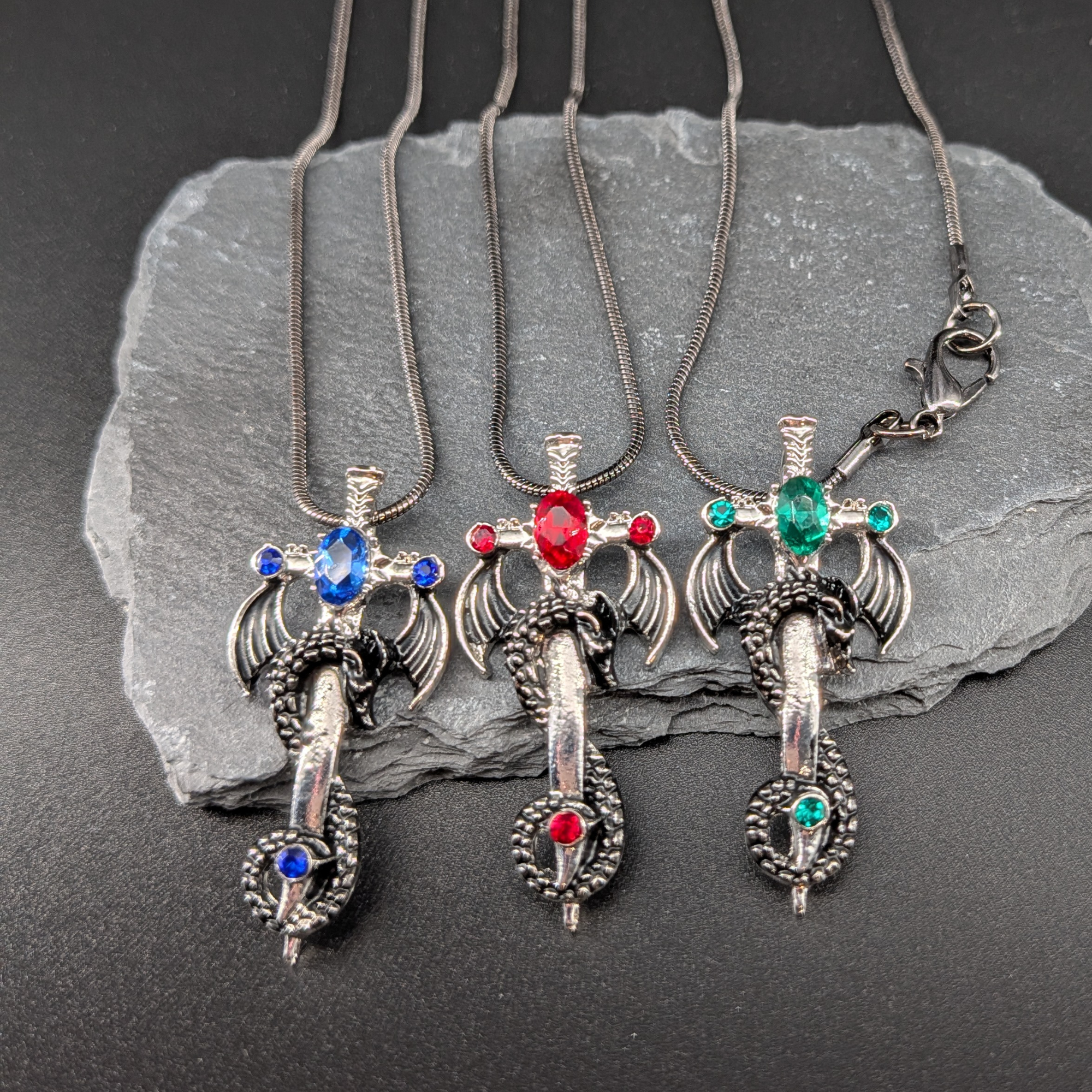 Three silver dragon necklaces with colored gemstones on a dark stone surface